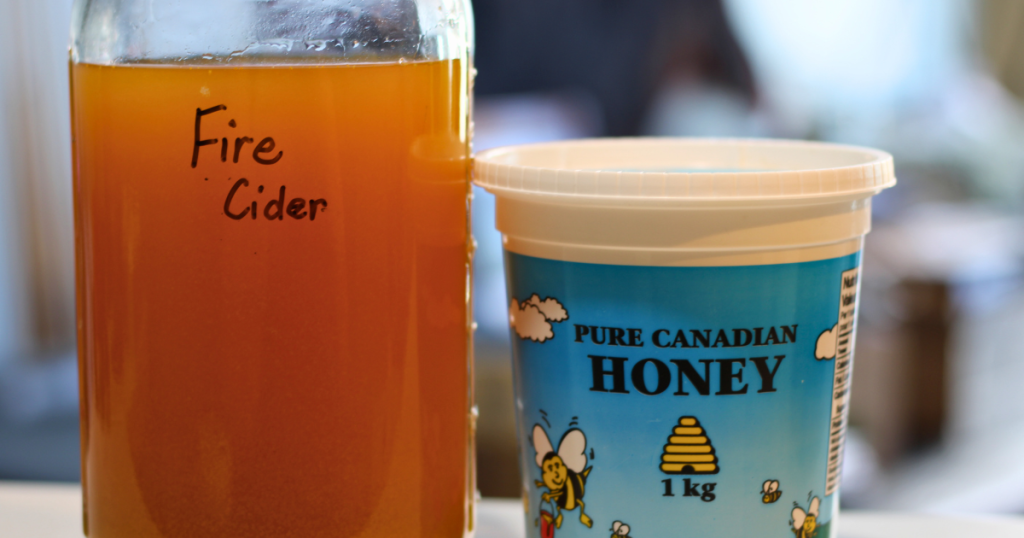 Strained fire cider in half gallon glass jar beside a container of pure Canadian raw honey.
