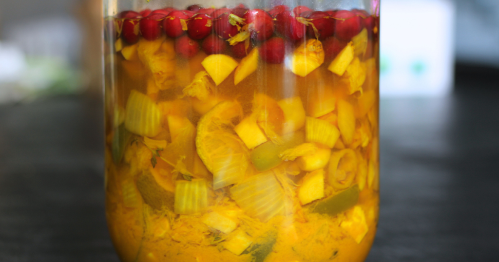 Fire cider ingredients, such as limes, onions, ginger, garlic and cranberries floating in a glass jar.