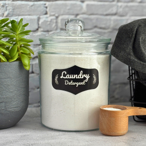 Homemade non-toxic laundry detergent in a glass container next to a green plant and a basket with laundry.