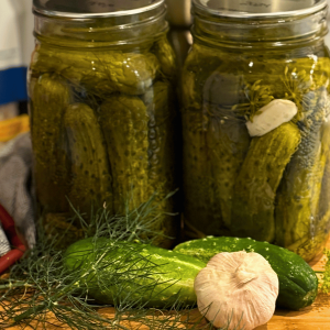 Two jars of dill pickles on the counter with cucumbers, garlic and dill.