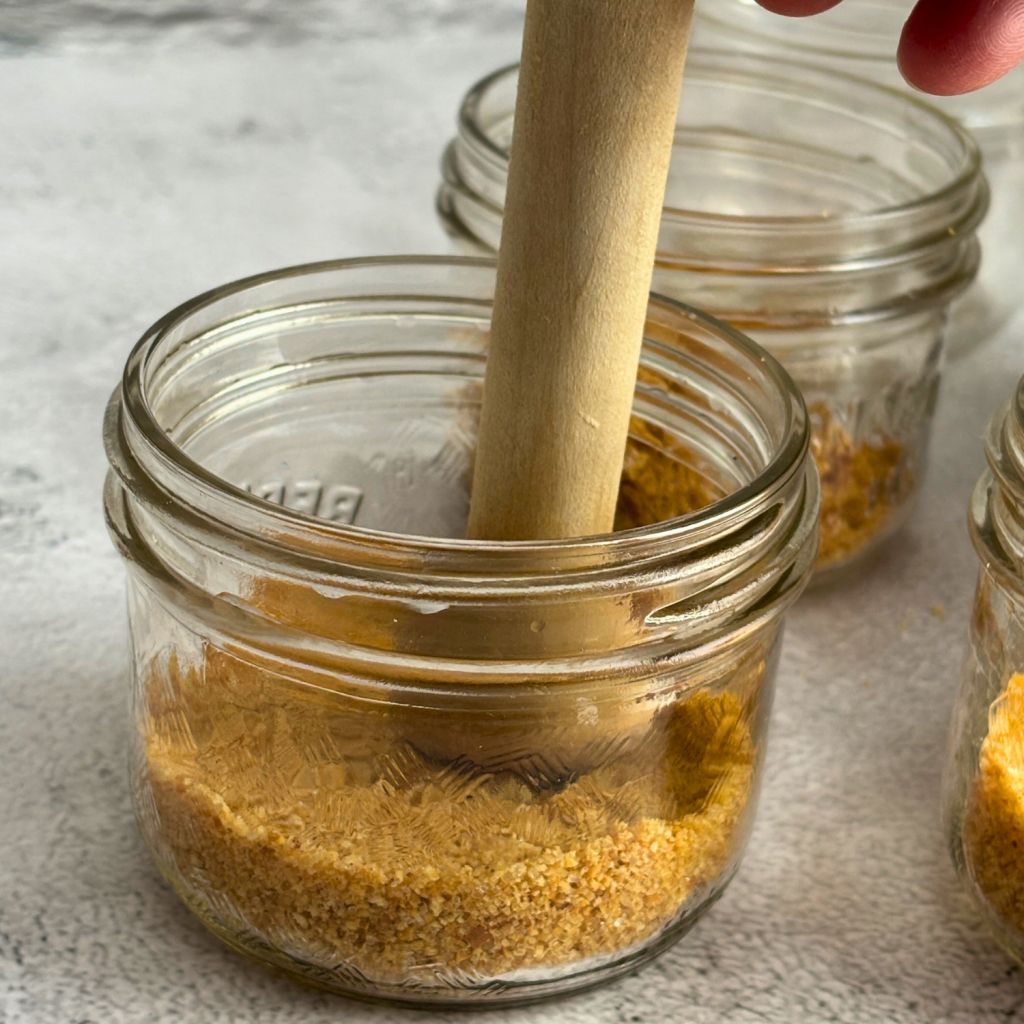Mason jar with tamping tools pushing the graham crust mixture down in the bottom of the jar.