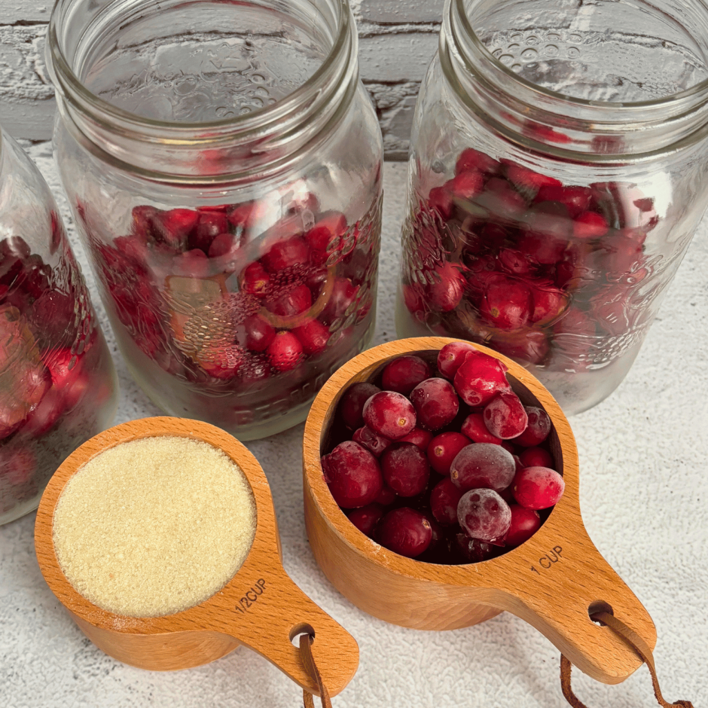 Prepping Three jars to can cranberries. Jar have cranberries in them and there is a  measuring cup with sugar and a measuring cup with cranberries.