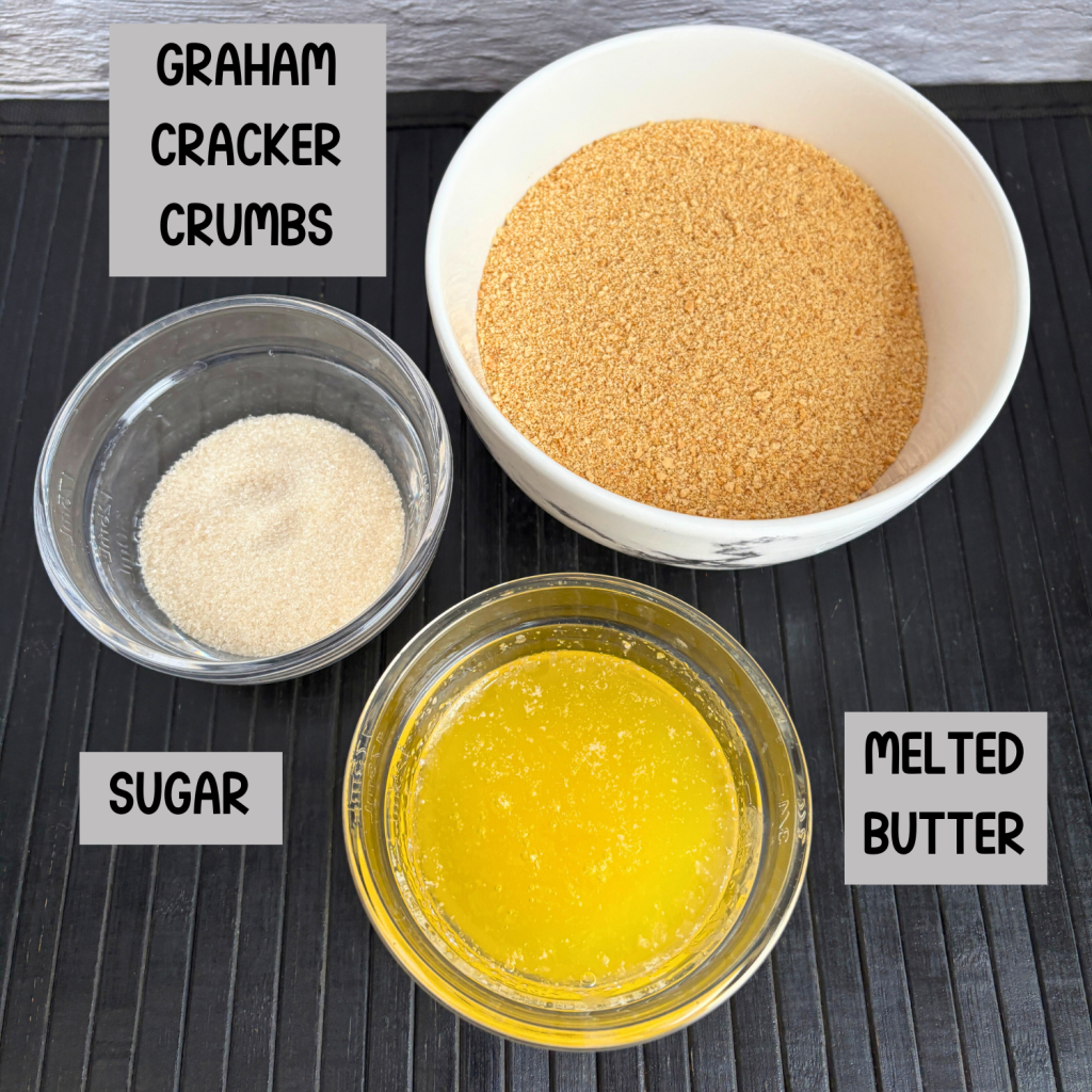 A bowl of melted butter, a bowl of graham cracker crumbs and a bowl of sugar on a black mat.