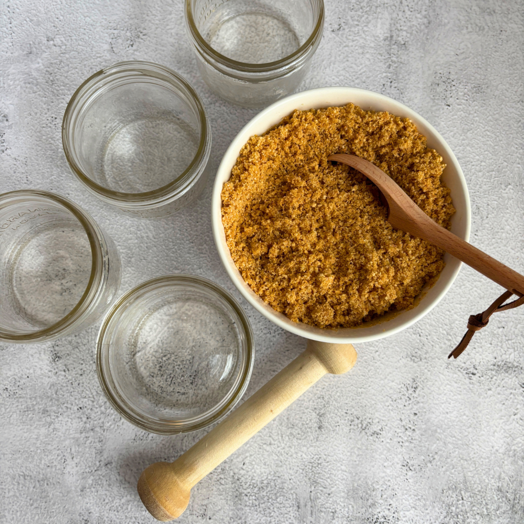 Bowl of graham cracker crust mixture, empty mason jars, a wooden spoon and a tamping tool.