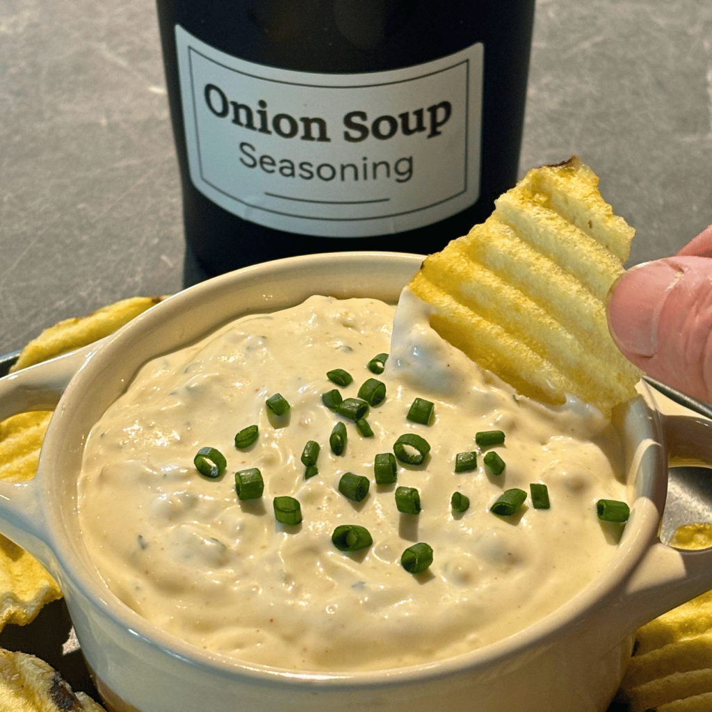 Bowl of homemade onion dip with a container of onion soup seasoning in the background and some dip on a ripple chip.