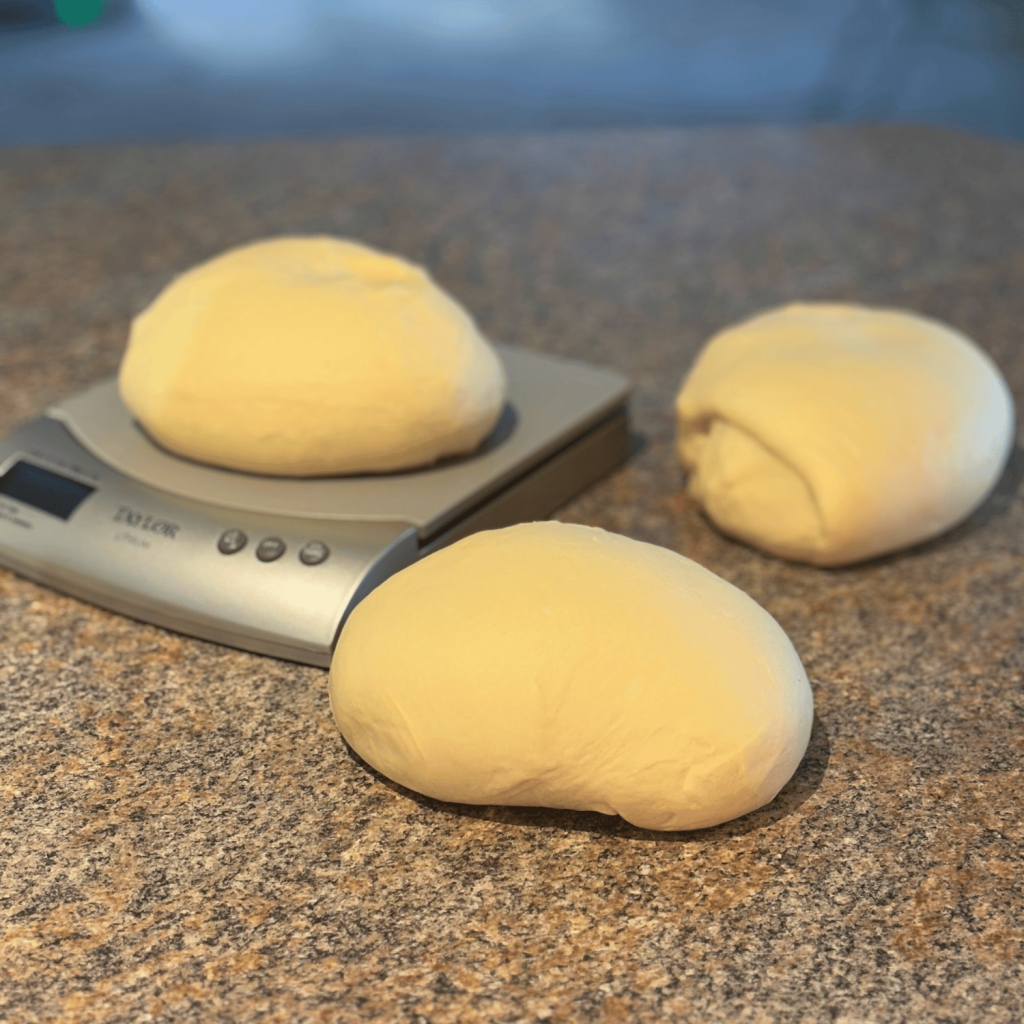 Three pieces of bread dough and one of them is on a kitchen scale.