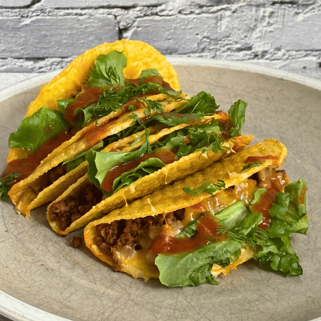 Plate with four beef tacos topped with cheese, lettuce and taco sauce.