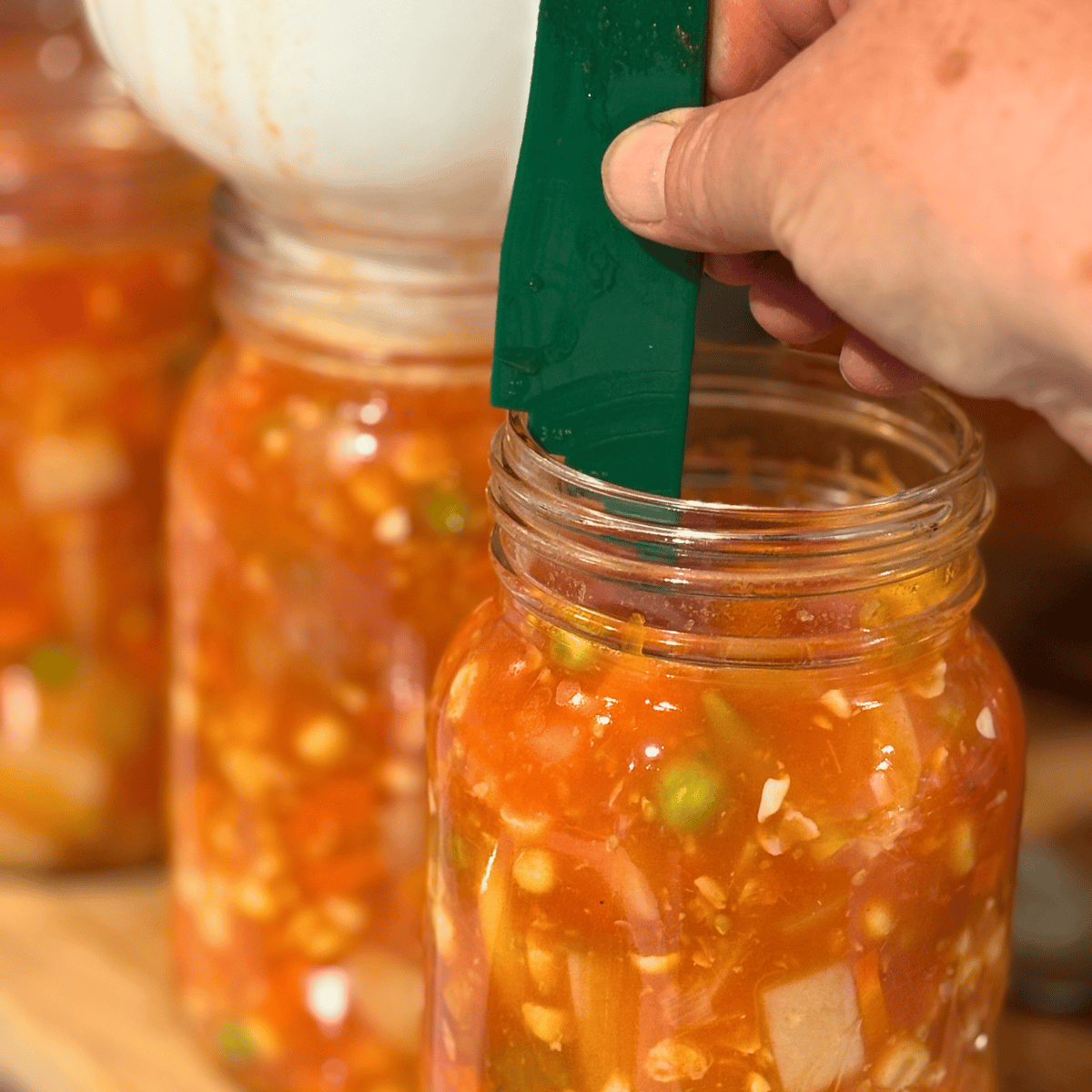 Three jars of homemade canned vegetable soup on a counter, one has a canned funnel in it and the other has a headspace canning tool.