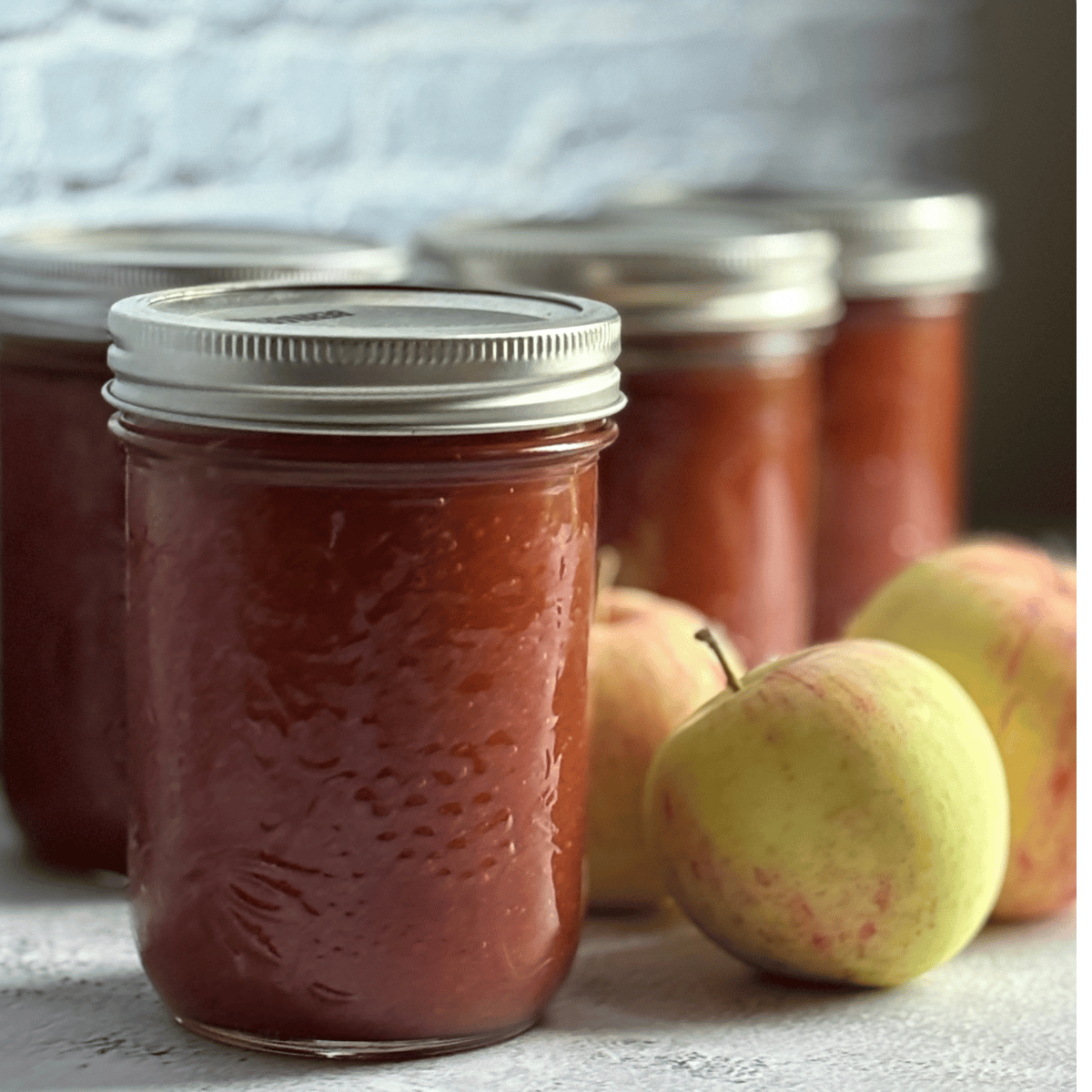 Jars of homemade apple sauce on a counter next to some apples.