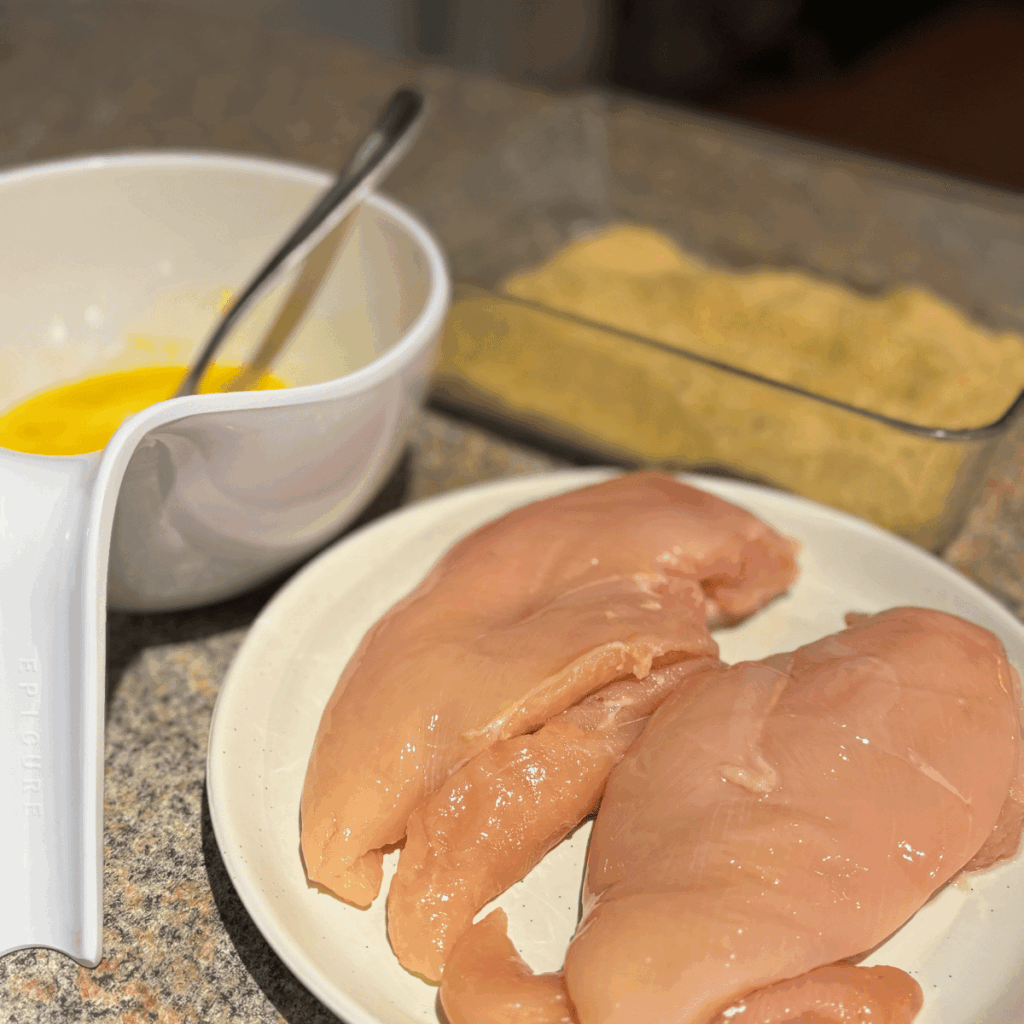 Breading station for chicken parmesan. Plate with chicken breast, bowl with eggs and a shallow dish with breadcrumbs