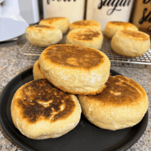 Black plate with homemade sourdough English muffins on it with some other English muffins in the background.
