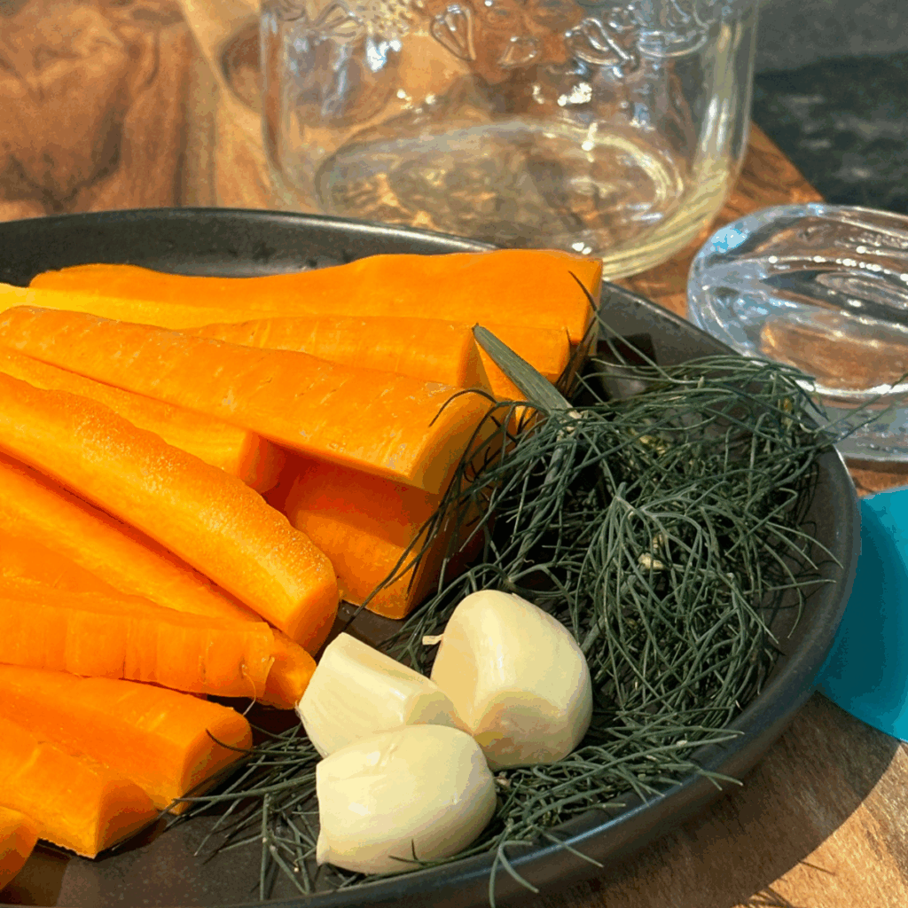 Ingredients for fermented carrots- carrot sticks, dill and garlic on a plate next to a quart jar and a glass fermentation weight.