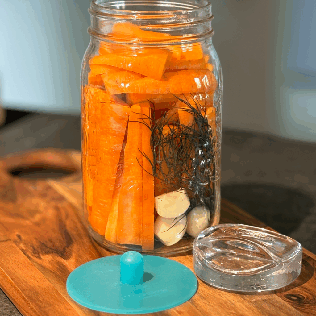 Quart jar filled with carrot sticks, dill and fresh garlic cloves on a cutting board next to a fermentation weight and a pickle pipe.