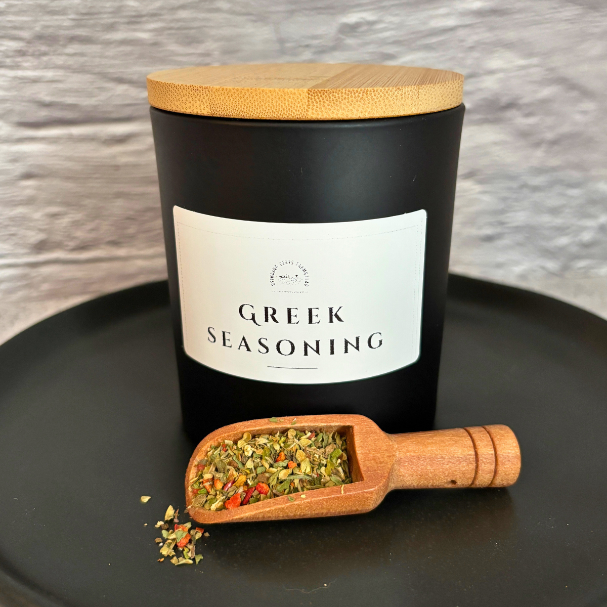 Container of homemade Greek seasoning on a black plate with a wooden scoop filled with the spice blend.